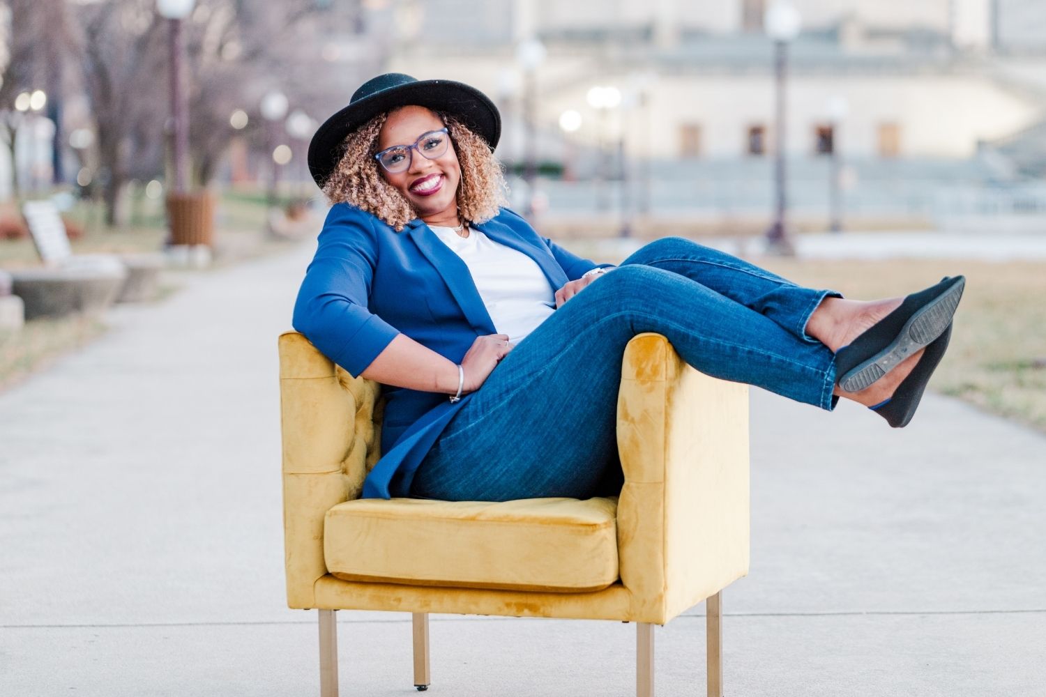 Woman in a black hat and blue blazer sitting in a yellow velvet chair outside with her legs dangled over, looking and smiling at the camera